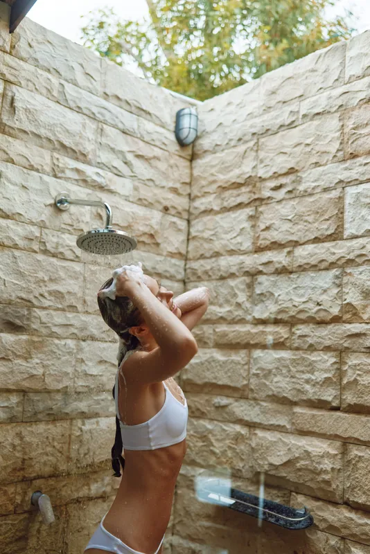 woman showering outdoors under rainfall, washing hair with soap, natural stone wall background, bright daylight, summer wellness routine