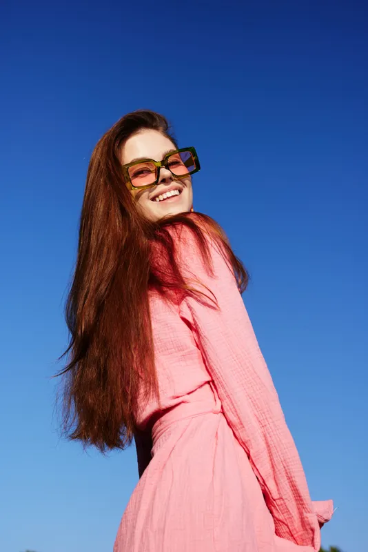 Young woman with long red hair smiling in a pink dress under a blue sky, showcasing vibrant summer fashion and joyful emotions