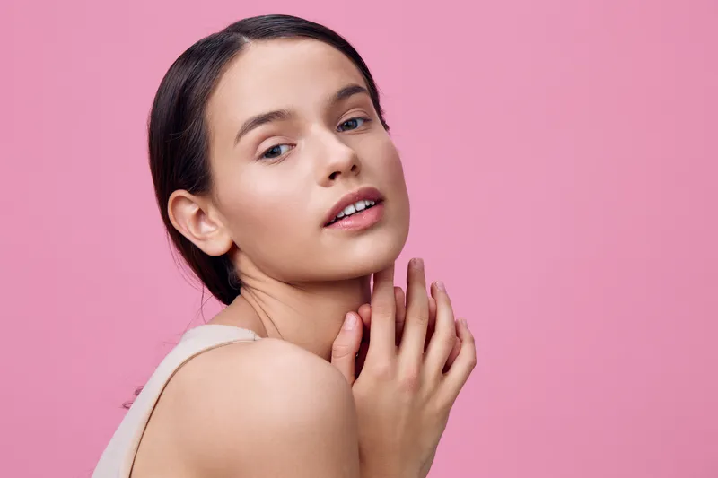 Portrait of a young woman with flawless skin and natural makeup against a pink background, conveying beauty and confidence in a fresh, modern style