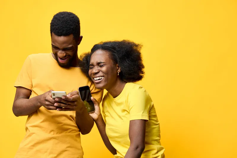 Joyful Black couple enjoying their time together with smartphones against a vibrant yellow background, showcasing happiness and connection