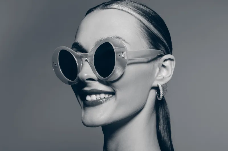 Smiling young white woman with light skin wearing large round sunglasses and hoop earrings. Portrait in grayscale with sleek hairstyle. Fashion and style concept.