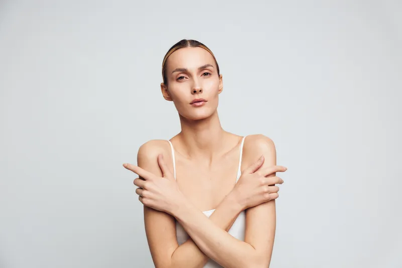 Young woman crosses her arms over her shoulders in a minimalist studio setting, wearing a white tank top, calm expression, and a clean light gray background, conveying confidence and poise.