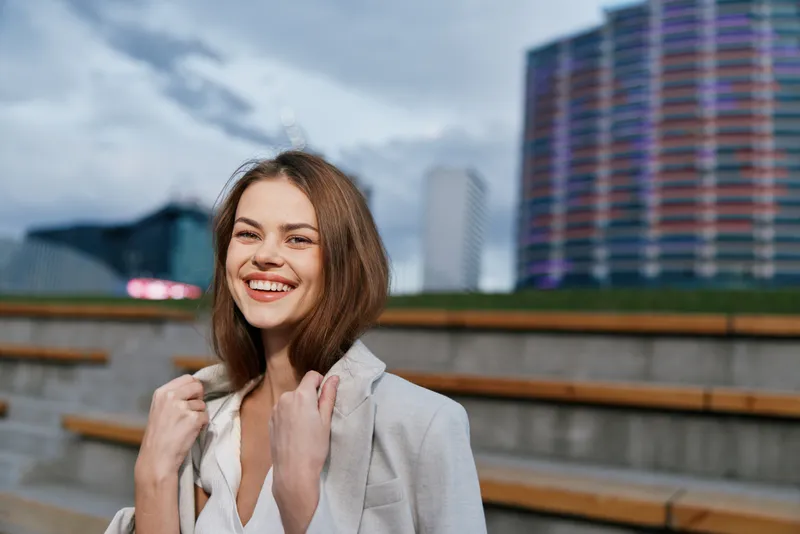 Confident woman in business attire smiling outdoors against a city skyline, showcasing professionalism and vibrancy in a modern urban environment.