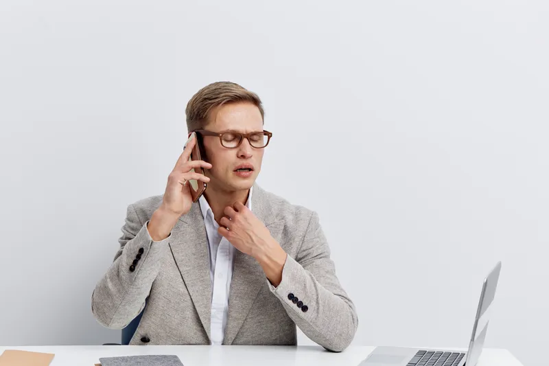 frustrated man touching throat talking on mobile phone wearing glasses grey jacket sitting at desk with laptop and notebook isolated on white background communication concept