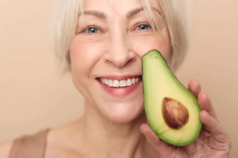 Smiling mature woman with light hair holds fresh avocado half near face against beige background. Concept of healthy lifestyle, natural nutrition, wellness, and beauty care for older adults.