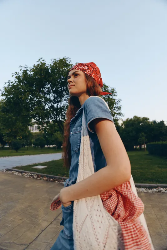 Lifestyle woman in modern boho-western denim street style walks outdoors with red bandana and tote bag, captured in warm film color tones under clear sky.