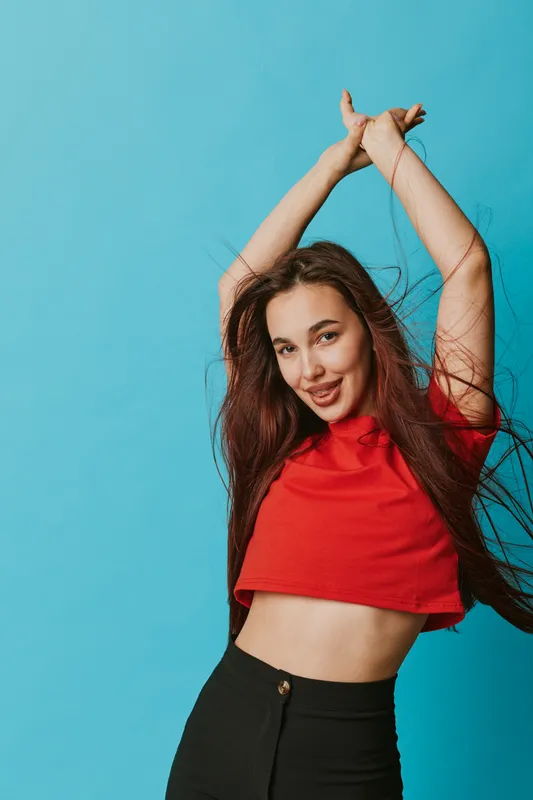 Young woman with long hair wearing red crop top and black pants posing with arms raised against blue background. Smiling female model with casual style and confident expression.