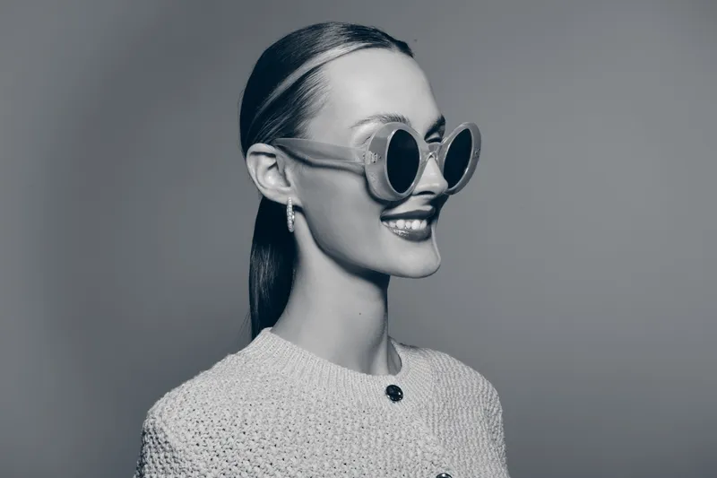 Smiling Caucasian woman with pale skin and slicked back hair wearing large round sunglasses and knitted sweater. Studio portrait with elegant and confident mood.