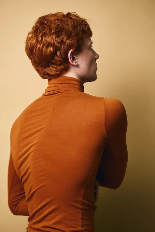 Woman with short red hair and orange turtleneck sweater posing with back turned against beige background in studio for fashion portrait and style concept.