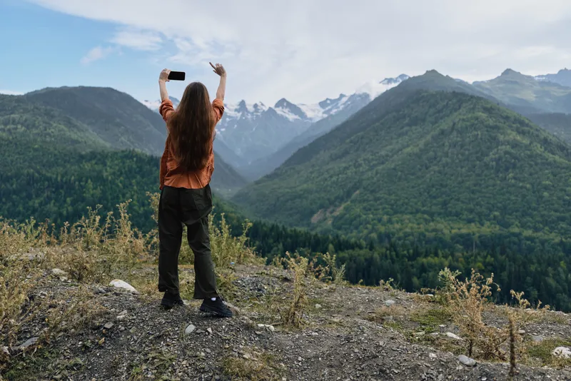 Hiker takes a selfie on a rocky overlook amid a dramatic mountain landscape, with lush valleys and evergreen forests, capturing outdoor adventure and expansive scenery