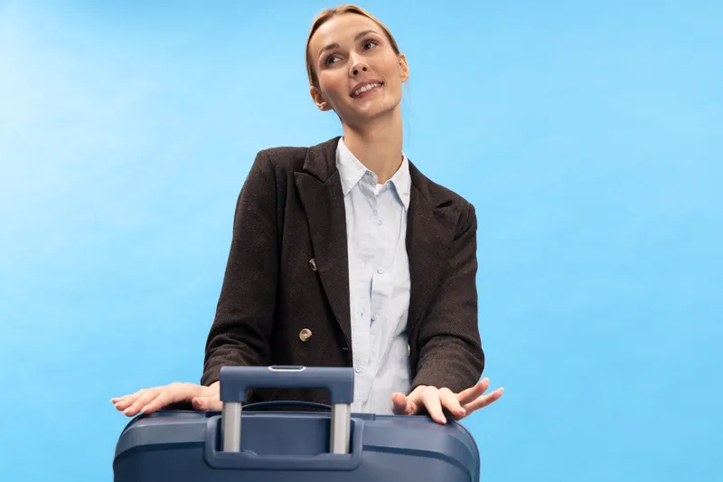 Young businesswoman in blazer stands with suitcase on blue background, looking away thoughtfully. Concept for travel, business trip, planning, and professional lifestyle.