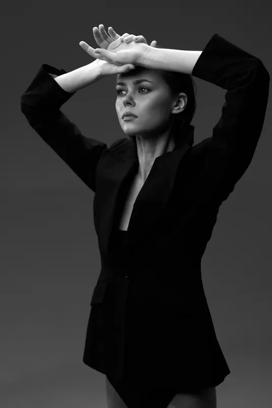 Black and white portrait of a serious woman in a black blazer with raised arms in studio light.