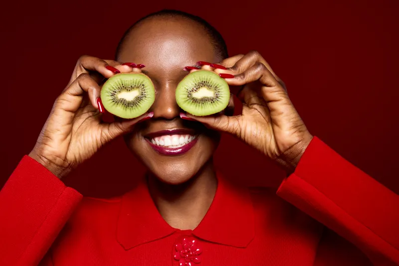 Smiling dark skinned African woman holds two kiwi halves over eyes in studio. Bright red outfit contrasts with brown background. Concept of healthy lifestyle, fruit, and joyful mood.