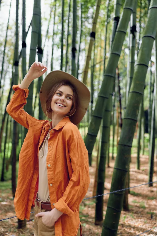 Woman in hat smiling in a bamboo forest portrait outdoors, wearing orange shirt and casual outfit, travel lifestyle, relaxed pose among tall green bamboo trunks and dappled sunlight filtered light