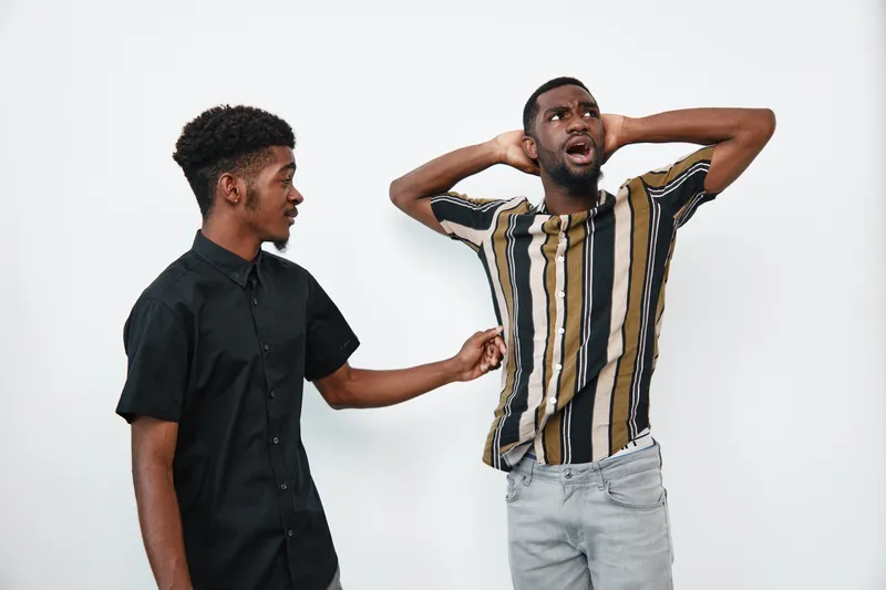 Two young black men with dark skin in casual clothes having a tense conversation on white background. One man looks surprised while the other gestures with concern and focus.