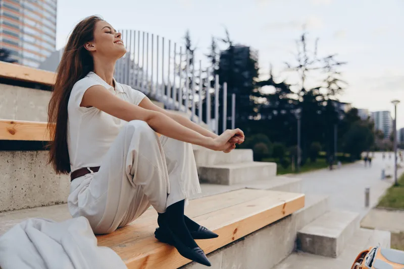 Young woman in white casual outfit sitting relaxed outdoors on wooden steps enjoying peaceful moment with city park background and soft natural light