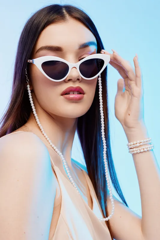 Young woman with long hair wearing white sunglasses and pearl accessories poses against a blue background. Concept of beauty, fashion, and modern style.