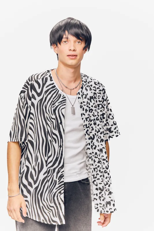 Young man wearing a black and white animal print shirt with layered necklaces stands against white background, showcasing modern casual fashion style and confident pose.