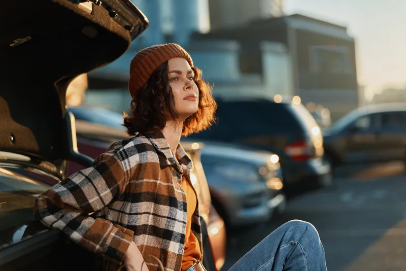 woman leaning on car trunk in parking, candid authenticity at golden hour with beanie and plaid jacket, relaxed lifestyle portrait capturing mindful living and emotional storytelling mood