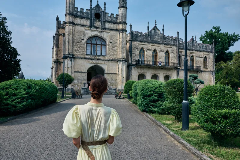 A woman in a vintage dress walks along a gravel path toward a grand historic castle, flanked by manicured hedges and classic lampposts under a bright blue sky today.