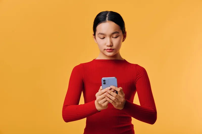 Young woman in red sweater using smartphone with concentrated face against yellow background for technology, communication, social media, connection, and online interaction concept.