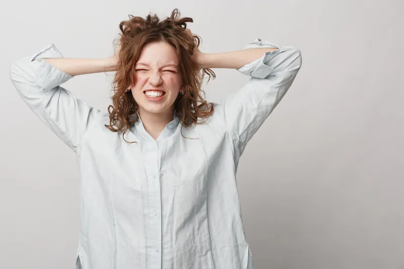 Frustrated young woman with curly hair wearing white shirt pulls her hair and grimaces. Isolated on light background. Concept of stress, anxiety, and emotional tension.