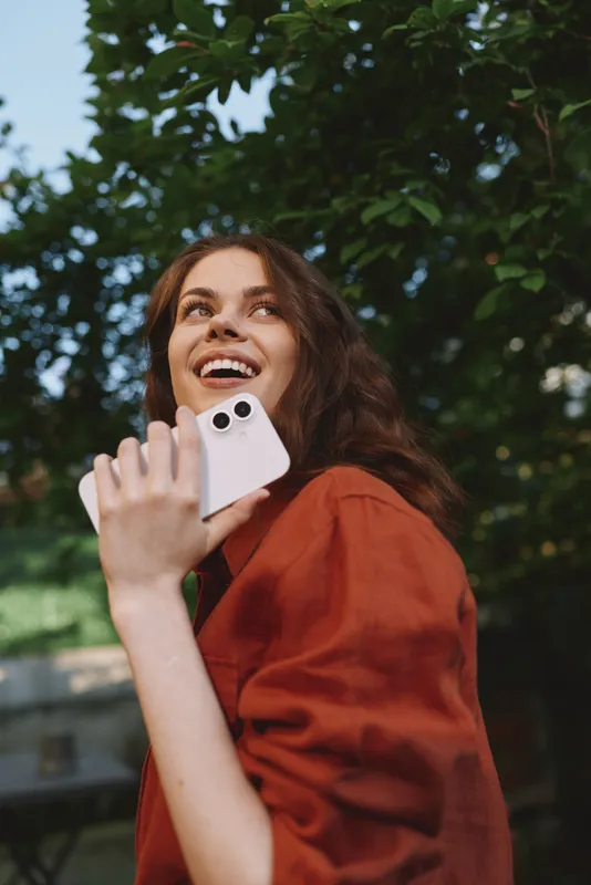 Smiling woman outdoors talking on a smartphone. Natural light and a leafy background create a warm, casual lifestyle mood