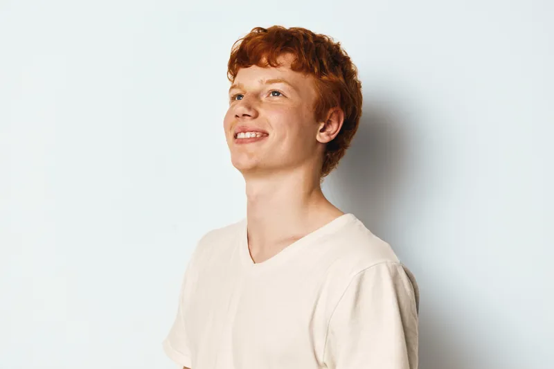 Young man with curly red hair smiling and looking up on white background. Casual male in white t-shirt posing with happy expression and relaxed posture in studio.