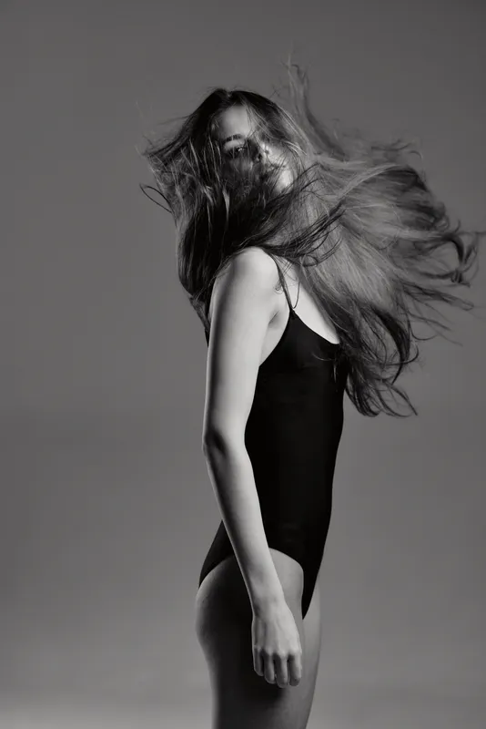 Black and white portrait of a young woman with flowing hair in a black bodysuit, showcasing motion and elegance.