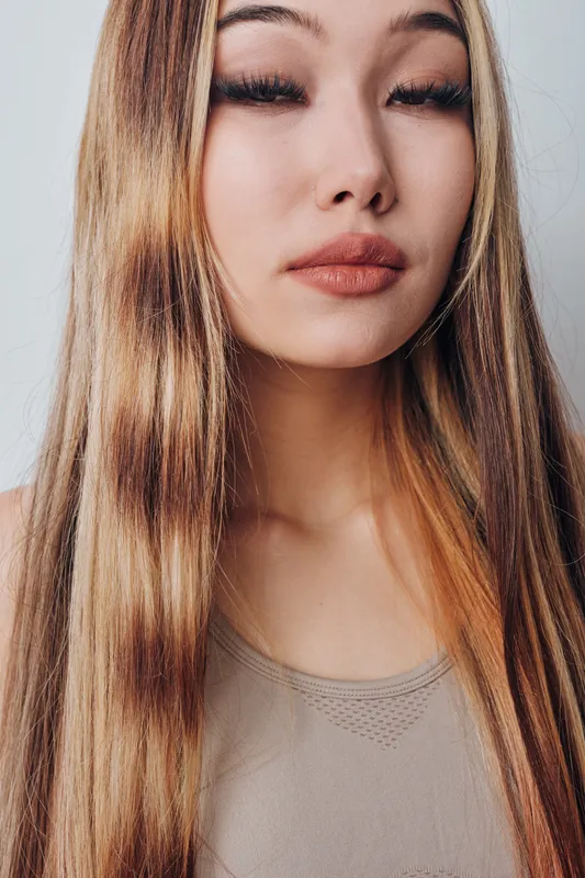 Young woman with long straight hair posing against white background, natural makeup, closed eyes and neutral expression in studio portrait with soft lighting and casual top.