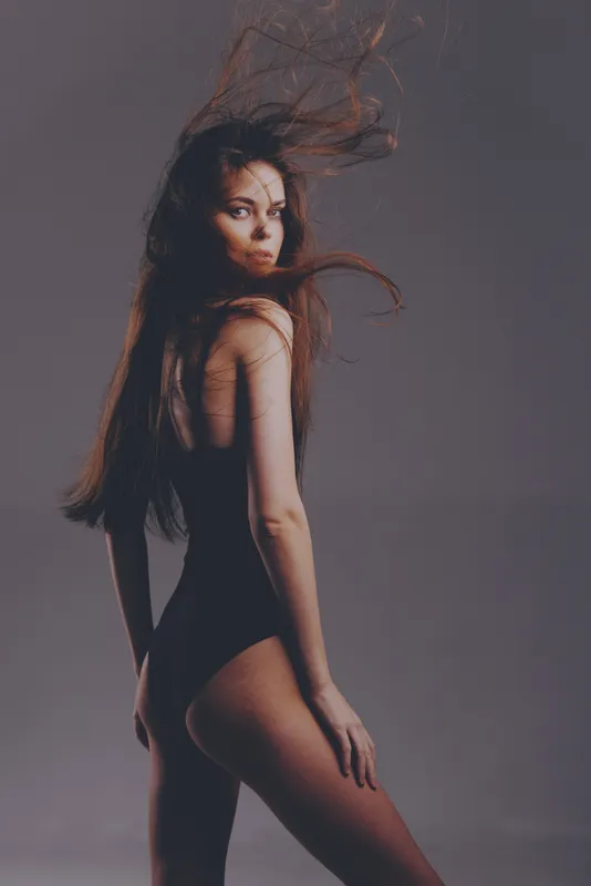 young woman in black bodysuit posing with flowing hair against gray background, fashion, elegance, beauty