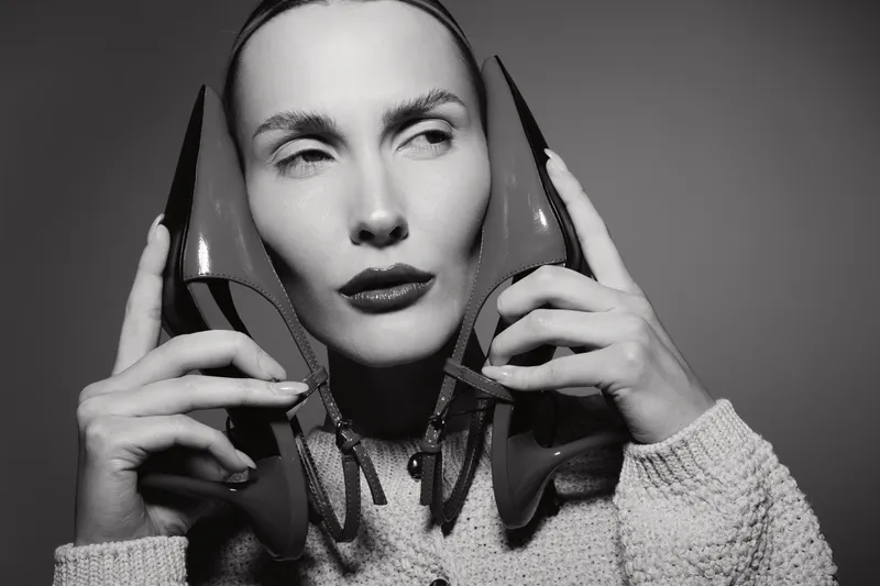 A fair skinned Caucasian woman holds red high heels near her face with a contemplative expression. Black and white studio portrait with soft lighting and fashion concept.