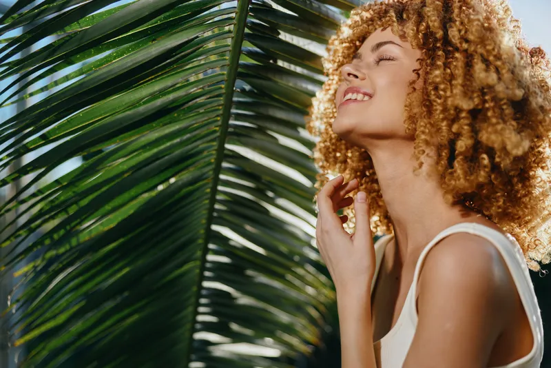 smiling woman enjoying tropical sunlight near palm leaf, curly hair, warm atmosphere, relaxed mood, summer vibes, natural beauty outdoors