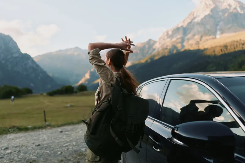 Young woman with a backpack stands beside a parked car in a valley, hands behind her head, gazing at distant mountains, enjoying a tranquil outdoor moment of freedom