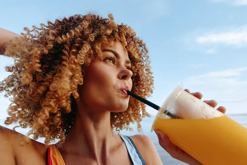 Woman with curly hair sips a drink through a straw, seaside summer vacation portrait outdoors, tropical beach breeze, holding an iced beverage glass, sunlit ocean sky, relax and smile warmly