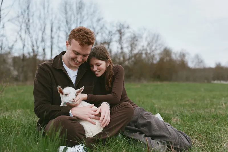 Lifestyle couple sits on a grassy field, smiling as they cuddle a young goat kid, enjoying a calm rural moment, casual clothes, soft natural light, peaceful affectionate scene in open park