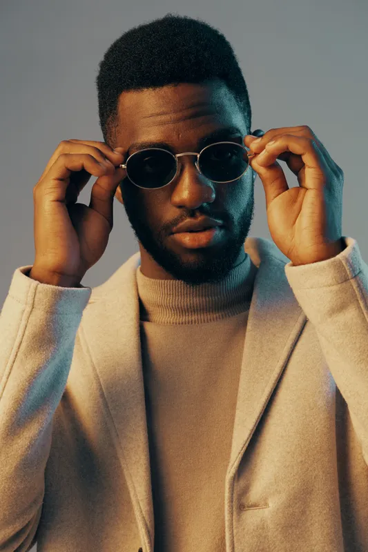 Stylish young man wearing sunglasses and a tailored blazer, showcasing confidence and sophistication against a soft pastel background