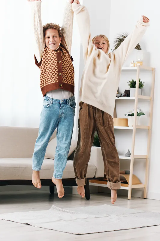 Authentic lifestyle image showing two children jumping in a cozy living space, capturing playful energy and simple joy while dressed for school days at home.