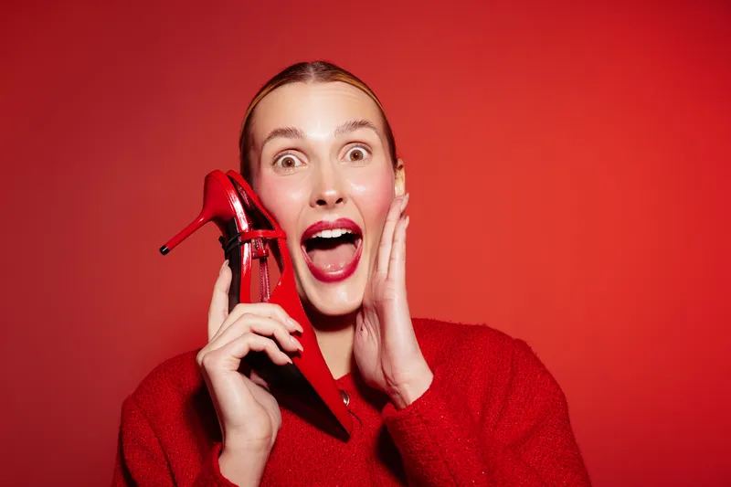 Surprised woman in red sweater holds red high heel shoe near face on red background. Concept of fashion, style, excitement, and vibrant color contrast in studio portrait.