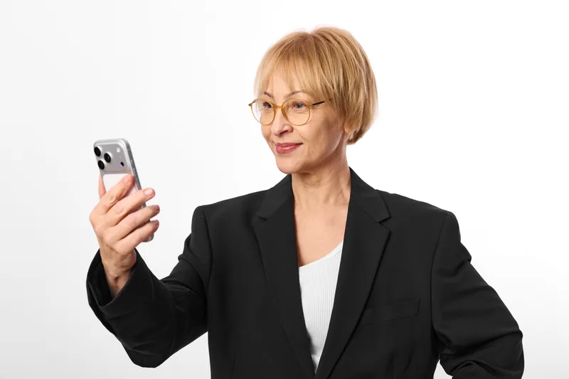 Smiling elderly woman with glasses holding smartphone, black blazer and white top, isolated on white background, using mobile communication, friendly technology concept, business support
