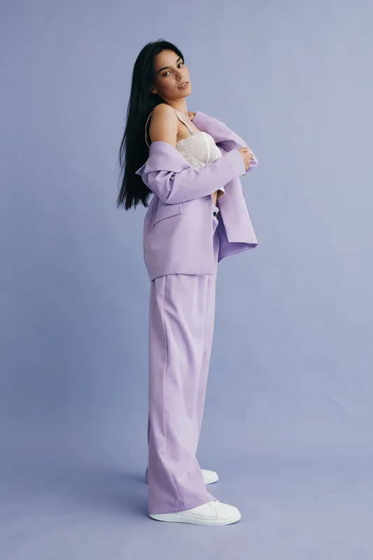 Stylish woman in lilac pantsuit with white sneakers posing confidently against soft gray background, showcasing modern fashion and relaxed elegance in studio.