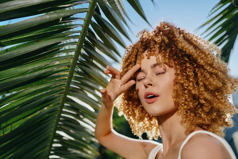 curly woman enjoying sunlight with closed eyes near tropical palm leaves in bright natural outdoor setting, capturing peaceful summer mood and fresh greenery