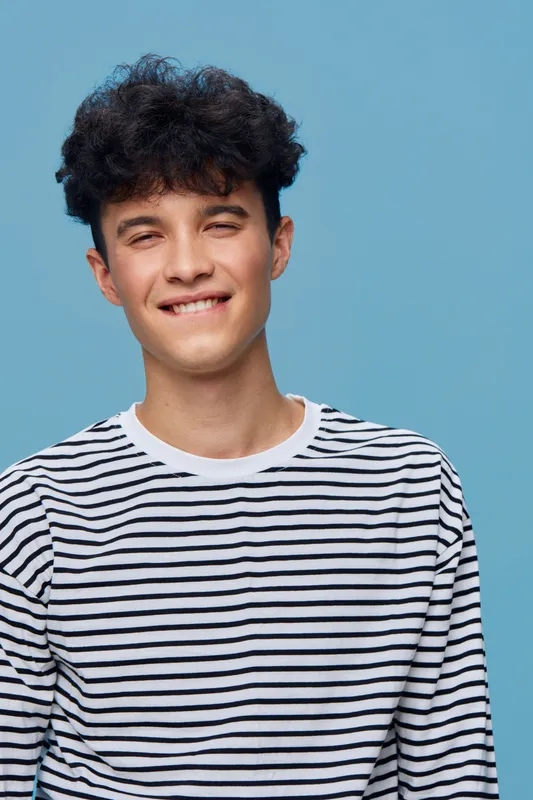 young man smiling wearing striped shirt with curly dark hair against blue background in casual style portrait