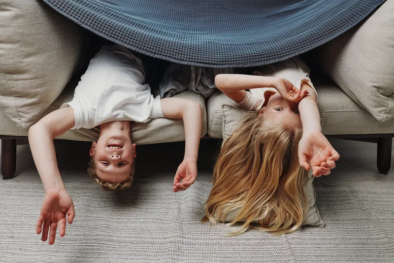 Two happy children lying upside down on a sofa covered with a blanket, smiling and playing in a cozy living room with neutral tones and soft natural light.