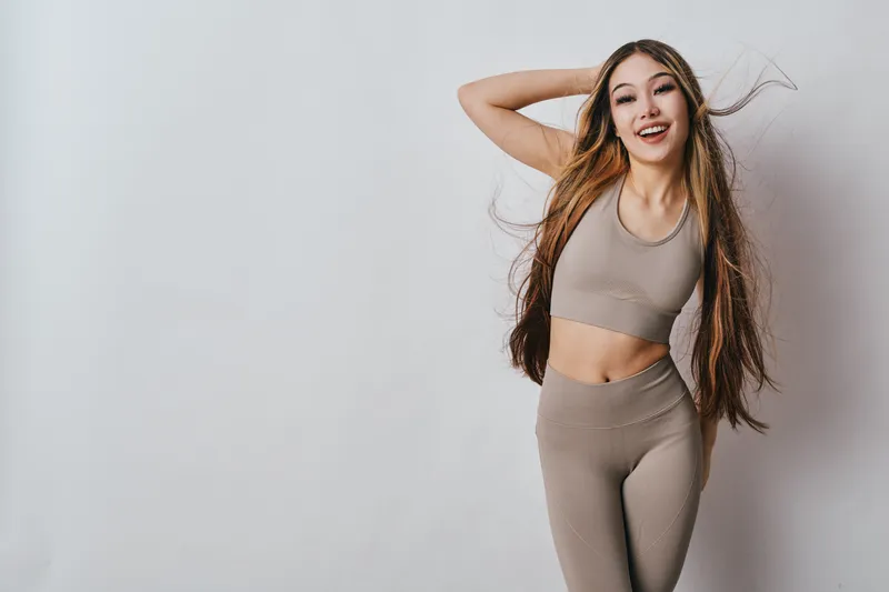 Young woman in beige sportswear standing barefoot near wall posing with hand behind head smiling, fitness model with long hair and athletic body for healthy lifestyle.