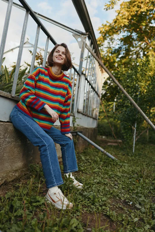 Woman in a rainbow sweater sitting outdoors on metal stairs, smiling and relaxed in casual jeans and white sneakers, leaning on railing in a green urban garden setting.