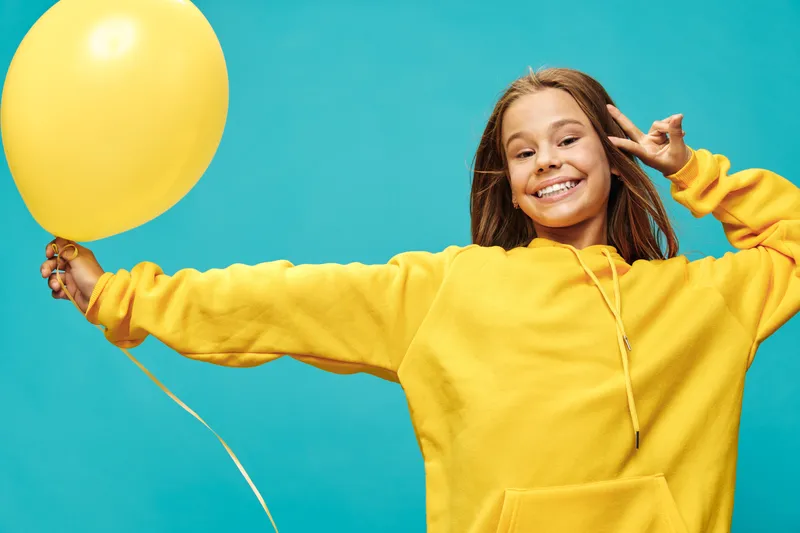 Smiling girl with long hair wearing yellow hoodie holds yellow balloon and shows peace sign with raised arm on blue background for happy childhood and celebration.