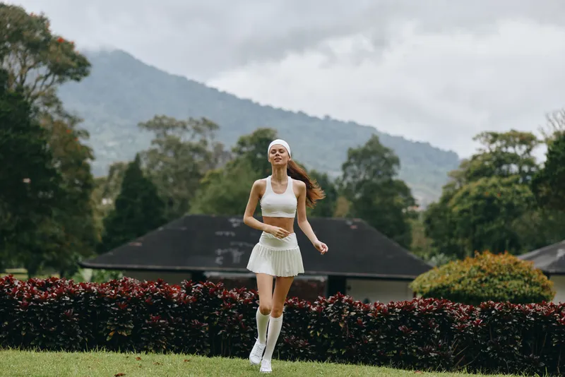 Young woman in white sports attire jogging joyfully in a lush green park with mountains in the background, emphasizing fitness and healthy lifestyle