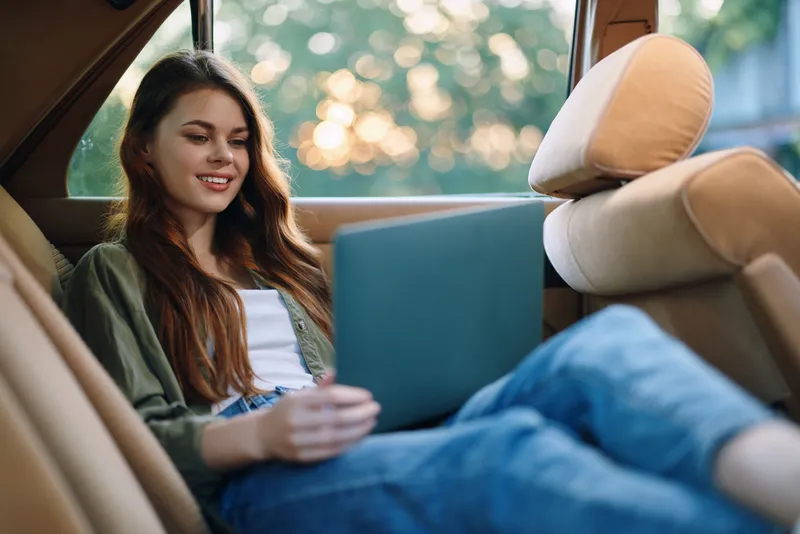 Young woman using a laptop in a cozy car setting, enjoying a relaxed moment with natural sunlight in the background, conveying a sense of comfort and connection.