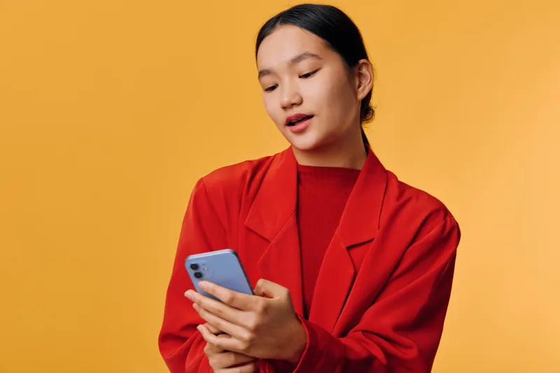Young woman in red jacket using smartphone with focused expression on orange background. Casual technology use, communication, and mobile interaction with device in modern style.